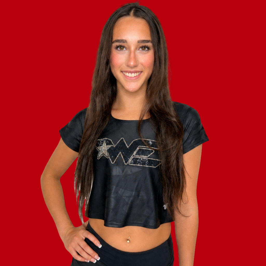 Young woman with long dark hair smiling at the camera, wearing a black cropped shirt with a silver star/diamond design, against a solid red background.
