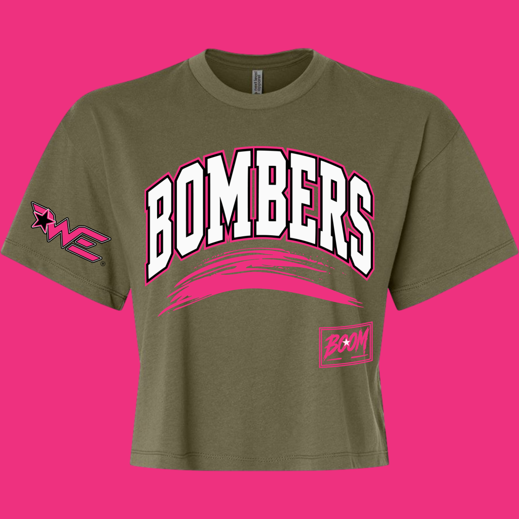 Olive T-shirt with a large white 'BOMBERS' arch logo outlined in pink and a pink brush stroke underneath against a pink backdrop.